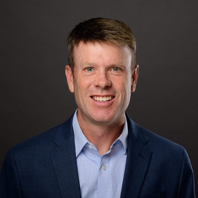 Headshot of Michael Oneill wearing a blue gingham dress shirt and navy blazer.
