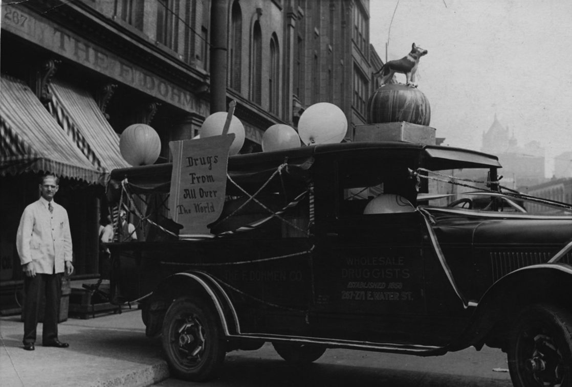 Black and white photo from the 1920s of a decorated Dohmen Company truck in a parade with balloons and a sign reading ‘Drugs from All Over the World,’ parked outside the storefront.