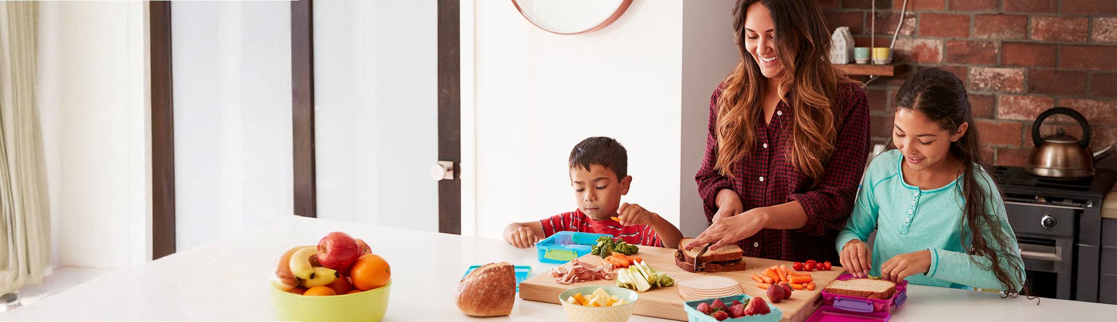 Smiling mother and two children making healthy lunches together in a bright kitchen. The boy is focused on packing his lunchbox while the girl slices bread next to her mother. Fresh fruits, vegetables, and sandwich ingredients are spread on the counter.