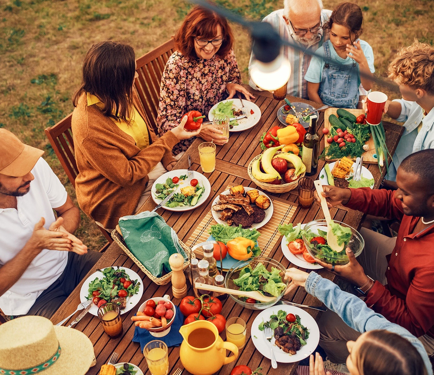 A diverse group of people enjoying a vibrant outdoor meal together at a wooden table filled with fresh fruits, vegetables, grilled foods, and salads. The setting suggests a warm, communal gathering with shared conversation and laughter.