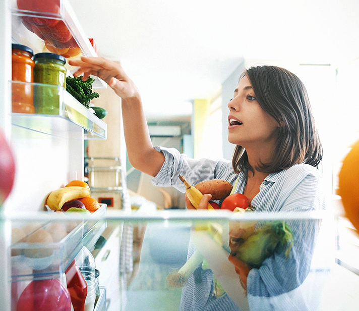 Young woman placing jars and produce on a refrigerator shelf, holding a variety of fresh fruits and vegetables in her arms, including bananas, tomatoes, and lettuce.