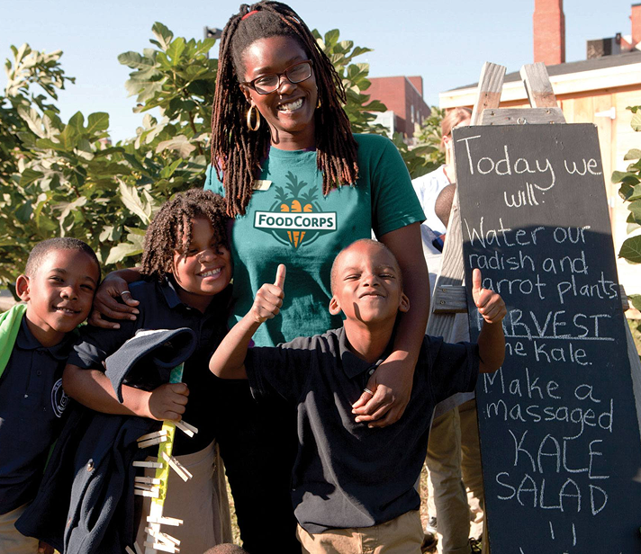 FoodCorps team member smiles with a group of elementary school children in a garden. A chalkboard lists activities like watering plants, harvesting kale, and making a massaged kale salad.”