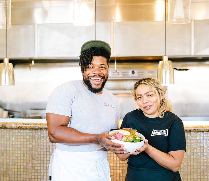 Two smiling restaurant staff members standing in a commercial kitchen, proudly holding a colorful bowl of fresh food.