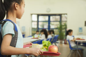 Young student with a backpack holding a red lunch tray containing an apple, salad, wrap, and drink in a school cafeteria, with other children seated in the background.