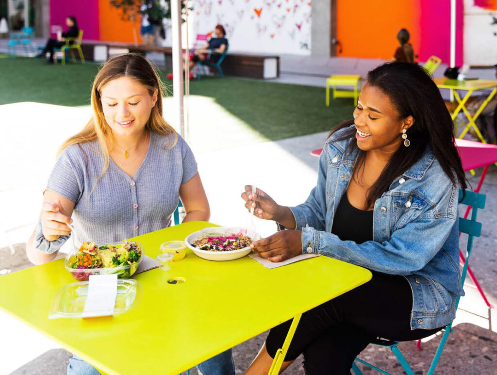 Two women sitting at a bright yellow table outdoors, smiling and enjoying fresh Everytable meals.