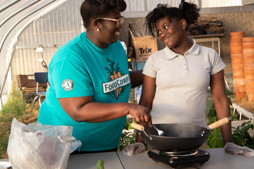 FoodCorps mentor teaching a student how to cook with fresh vegetables in a greenhouse setting.