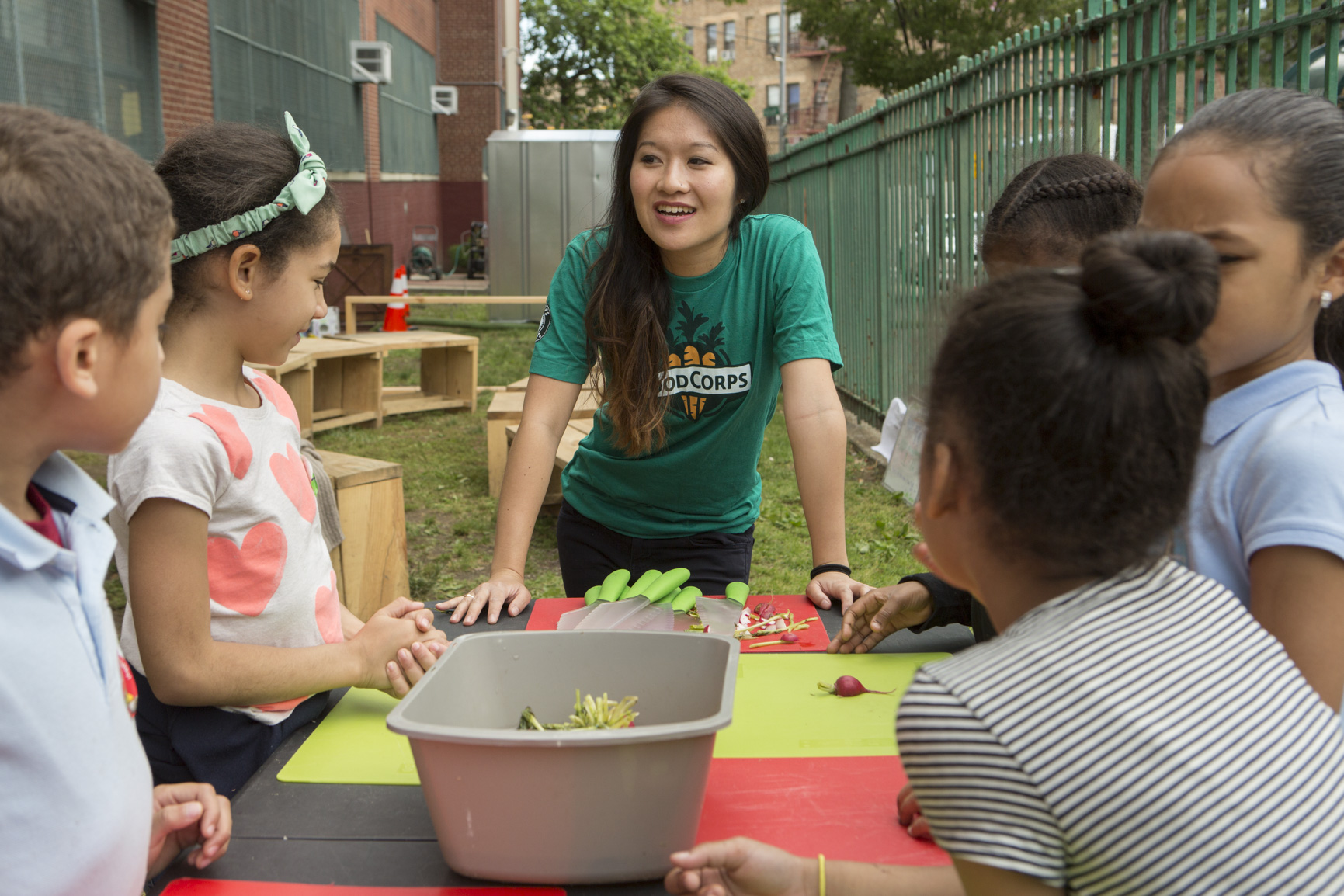 FoodCorps educator in a green T-shirt engages a group of elementary school children around an outdoor table in a Bronx school garden, preparing vegetables together.”