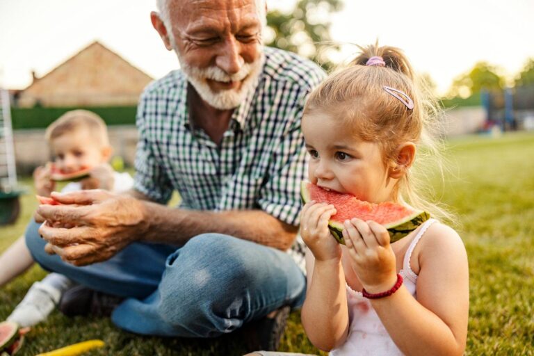 Smiling grandfather sitting on grass with his young granddaughter, who is happily eating a slice of watermelon on a sunny summer day.